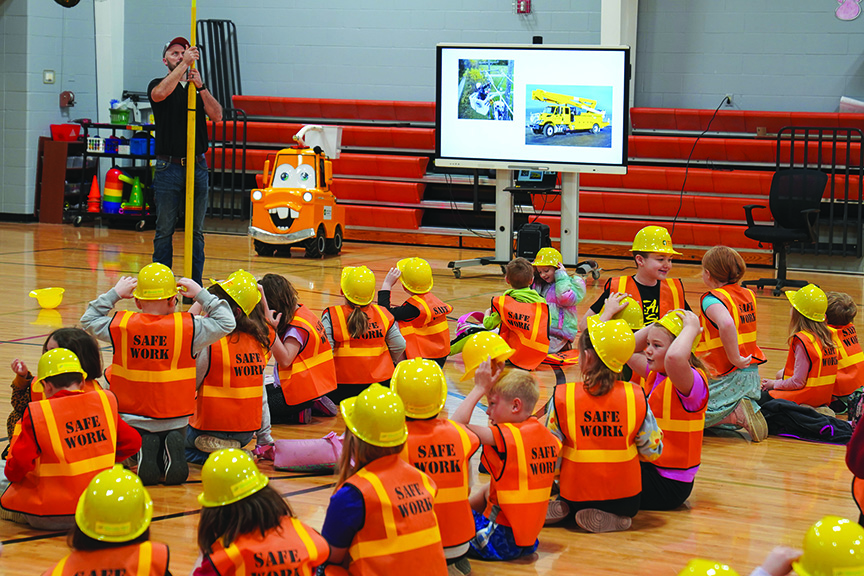 Power worker provides demonstration to several schoolchildren wearing safety vests and hats