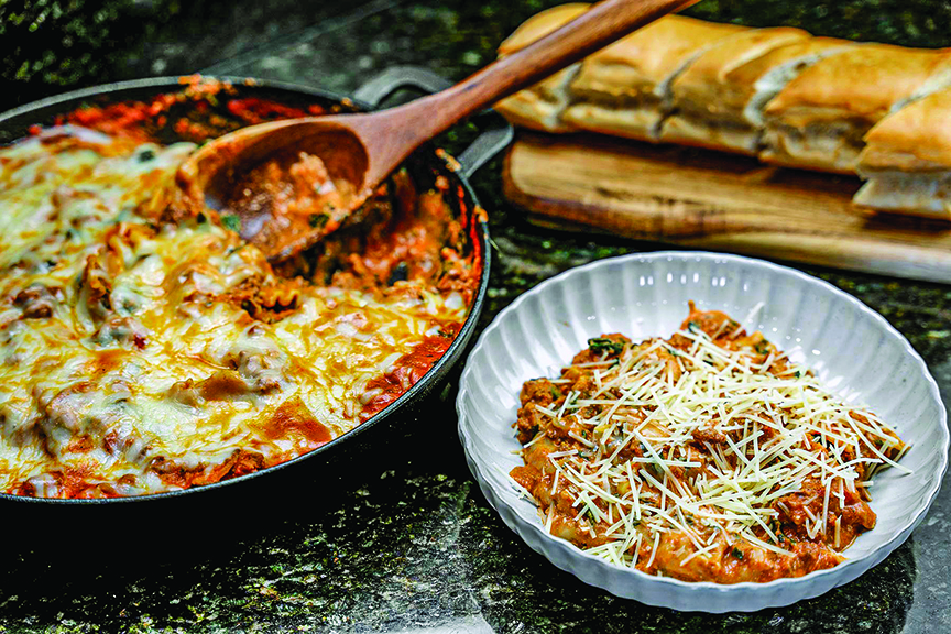 A skillet containing lasagna next to plate containing a serving near a loaf od bread