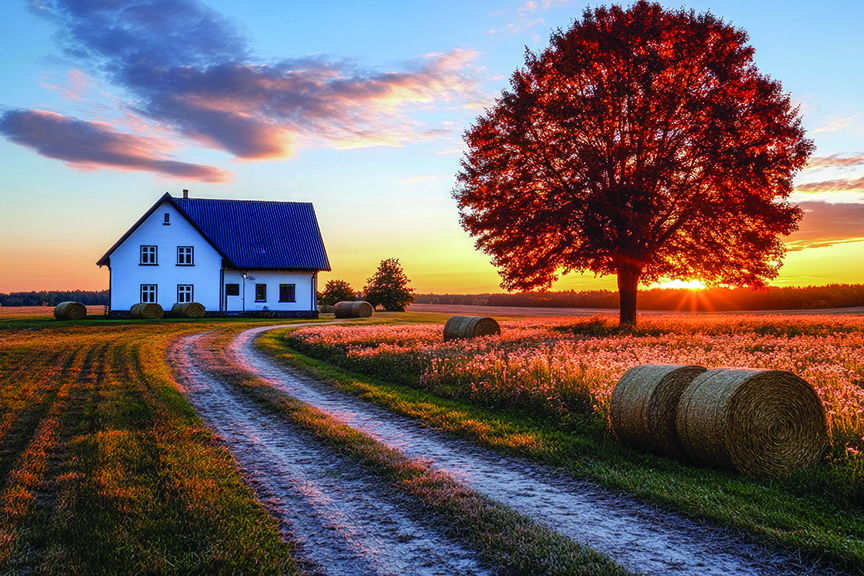Picturesque farmhouse at sunset in a tranquil rural setting. Hay bales, tree, and a dirt road complete the scene.