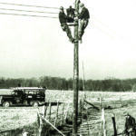 Black-and-white image shows linemen at work in a rural setting