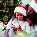 Mother hugs daughter in front of Christmas tree as daughter holds present. Both are wearing santa hats