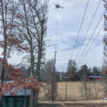 A helicopter flying above rural power lines, carrying a set of spinning saw blades suspended from a long cable.