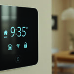 Close-up shot of a smart home automation control panel mounted on a light beige wall, showcasing a sleek design with rounded corners and a glossy black screen. The screen displays various icons and a digital clock reading 9:35 in bright blue color. Soft, diffused lighting creates a modern and comfortable ambiance. In the blurred background, a glimpse of an interior space with a wooden table and a window.