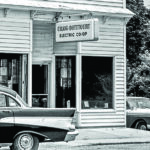 A black-and-white photograph shows the storefront of the "Craig-Botetourt Electric Co-op." The building features white horizontal siding and large display windows. A sign hanging above the entrance displays the name of the cooperative. Two vintage cars are parked on the street in front of the building.