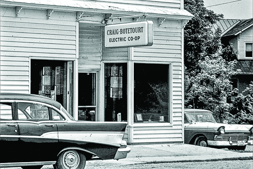 A black-and-white photograph shows the storefront of the "Craig-Botetourt Electric Co-op." The building features white horizontal siding and large display windows. A sign hanging above the entrance displays the name of the cooperative. Two vintage cars are parked on the street in front of the building.