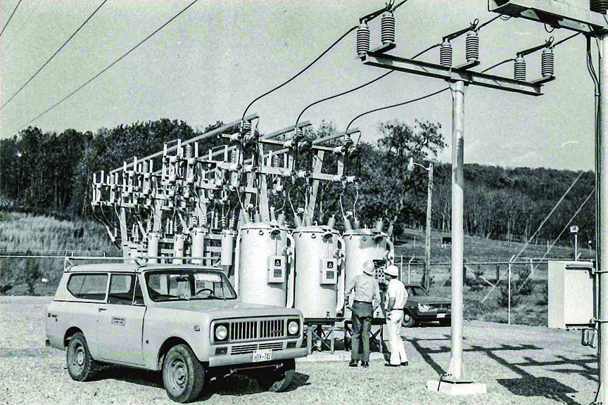 A black-and-white photograph shows two utility workers standing near three electrical transformers inside a fenced-in substation. A light-colored, vintage SUV is parked in the foreground on a gravel surface. Overhead, power lines connect to the transformers.