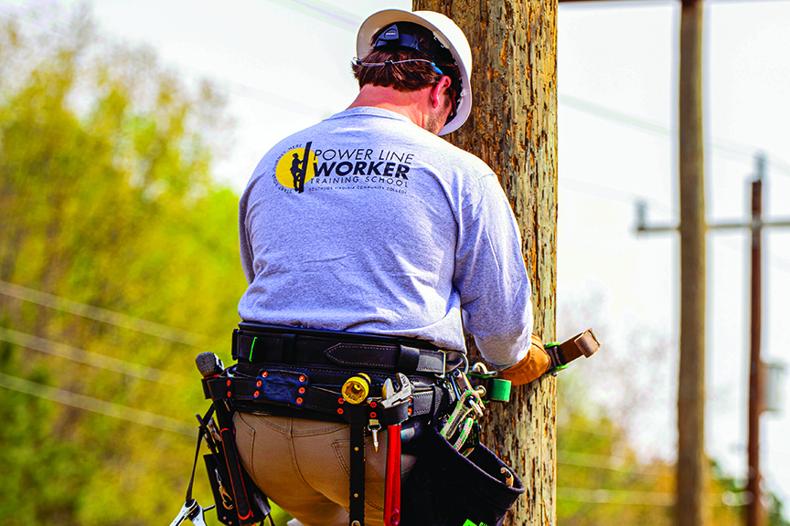 A rear view shows a power line worker wearing a white hard hat and a long-sleeved gray t-shirt that says "POWER LINE WORKER TRAINING SCHOOL" with a logo. The worker is leaning against a wooden utility pole while working, with a heavy-duty tool belt equipped with various tools fastened around their waist.