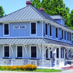 A two-story building in Paint Bank, Virginia, features light gray wood siding, a dark metal roof, and a wrap-around porch with a white railing. The structure has the words "PAINT BANK" displayed on a sign near the roofline, and the ground level is decorated with a row of vibrant orange flowers.