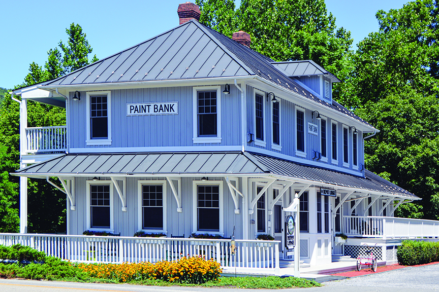 A two-story building in Paint Bank, Virginia, features light gray wood siding, a dark metal roof, and a wrap-around porch with a white railing. The structure has the words "PAINT BANK" displayed on a sign near the roofline, and the ground level is decorated with a row of vibrant orange flowers.