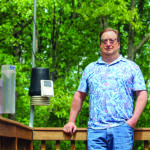 A man with short brown hair and glasses stands outdoors on a deck, smiling at the camera. He is wearing a short-sleeved Hawaiian shirt and blue jeans. At left, mounted on the deck railing and a metal pole, are weather-monitoring tools.