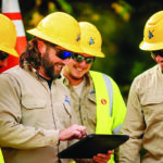 Four utility workers wearing yellow hard hats and safety glasses gather outdoors, smiling as they look at a tablet held by one of the men. They are wearing khaki work shirts and high-visibility safety vests. The logos on their hard hats and shirts read "Rappahannock Electric Cooperative."
