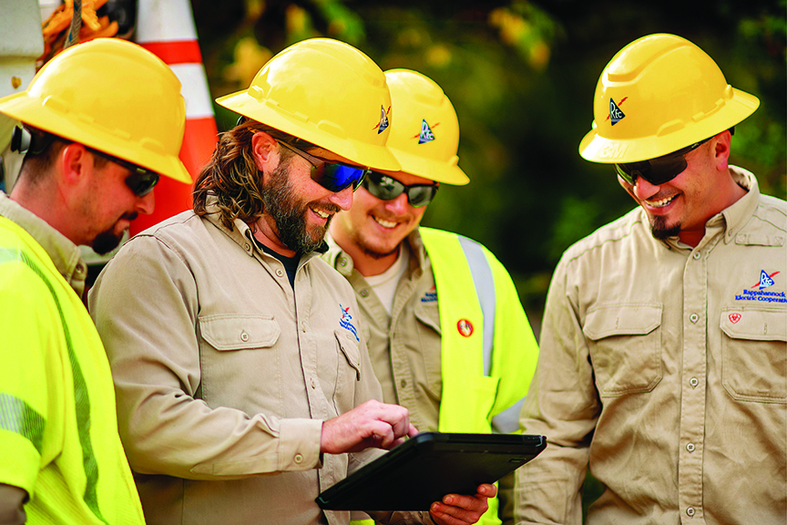 Four utility workers wearing yellow hard hats and safety glasses gather outdoors, smiling as they look at a tablet held by one of the men. They are wearing khaki work shirts and high-visibility safety vests. The logos on their hard hats and shirts read "Rappahannock Electric Cooperative."