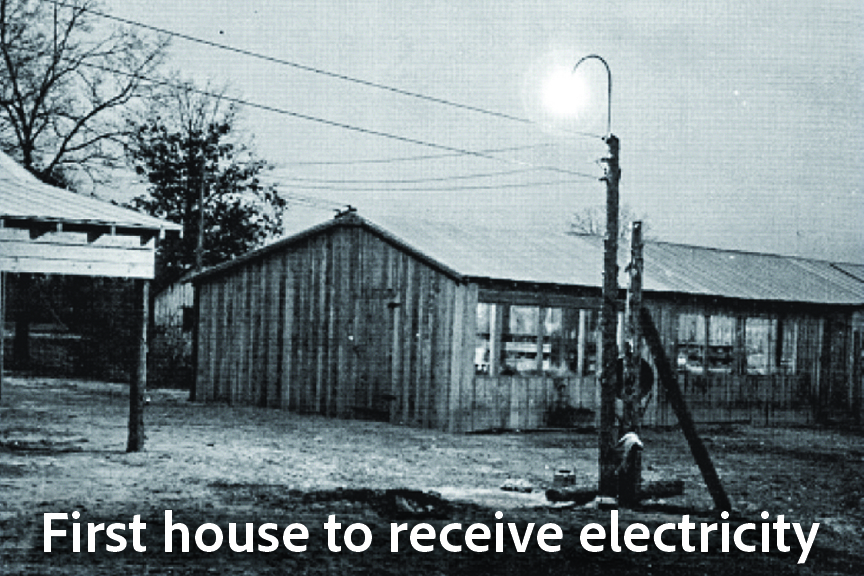 A historical, black-and-white photograph of a simple wooden farm building at dusk or night. An outdoor light fixture mounted on a tall wooden pole glows brightly, casting light over the ground. Power lines run horizontally across the top of the frame. White text at the bottom reads, "First house to receive electricity."