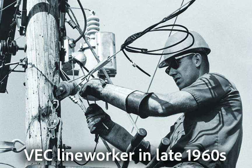 A late 1960s black-and-white photograph of a lineman working on a wooden utility pole. The worker, wearing a hard hat, safety glasses, and heavy protective gloves with long gauntlets, is manipulating power lines and hardware near a pole-mounted transformer.