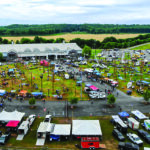 An elevated, wide-angle shot of a large outdoor lineman rodeo event on a grassy field next to a large pavilion building. The field is filled with dozens of tall wooden utility poles erected for climbing competitions, surrounded by crowds of people, vendor tents, and utility trucks. The foreground features parked pickup trucks, trailers, and white event tents, while a rural landscape of fields and forests stretches into the background under an overcast sky.
