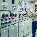 A smiling worker wearing a hard hat, safety glasses, and a plaid shirt stands in front of a switchgear wall at ODEC’s Wildcat Point Facility.