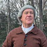 Andrew Packett, wearing a gray powdered wig, a white cravat, and a brown buttoned coat, stands outdoors against a background of bare trees.