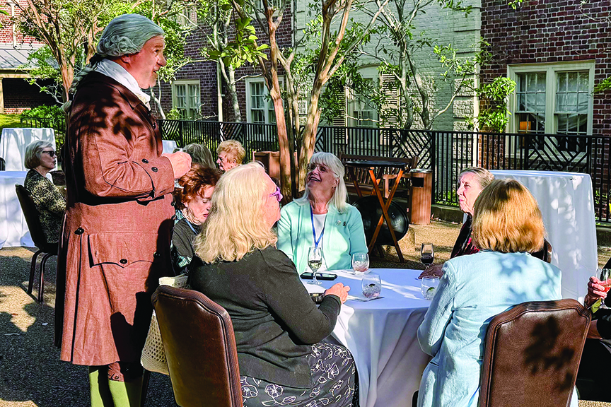 Andrew Packett, dressed in 18th-century historical attire, stands and speaks to a group of women with wine glasses seated around a round patio table.