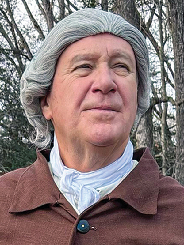 Andrew Packett, wearing a grey powdered wig, a white cravat, and a brown buttoned coat, stands outdoors against a background of bare trees.