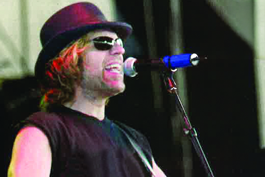 A close-up profile shot of a Kenny Alphin singing into a microphone on a stage. He is wearing a dark t-shirt, sunglasses, and a maroon fedora hat.