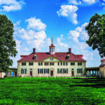 West front view of George Washington's Mount Vernon mansion under a bright blue sky with fluffy white clouds.
