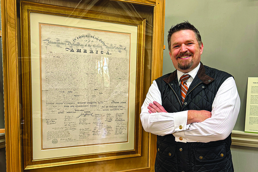 Man poses next to framed, historic copy of the Declaration of Independence