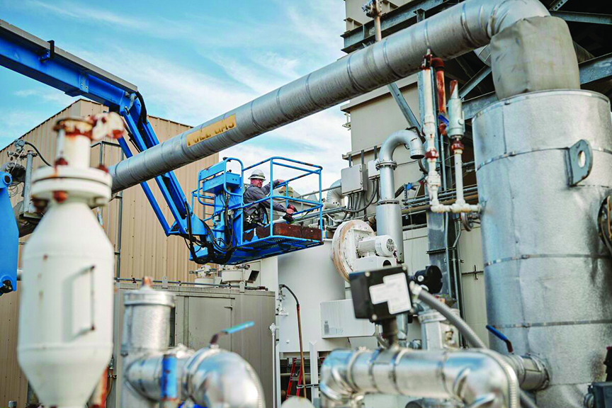 A facility technician working from an elevated blue boom lift surrounded by large industrial piping.
