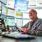 Man at desk surrounded by monitors smiles and looks up from a clipboard