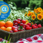 A woven basket filled with oranges, grapes, strawberries, and other fruits sits on a checkered blanket among wildflowers, creating a vibrant and inviting outdoor picnic setting.