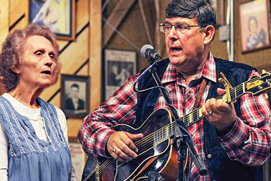 A man plays an acoustic guitar and sings into a microphone alongside a woman singing next to him.