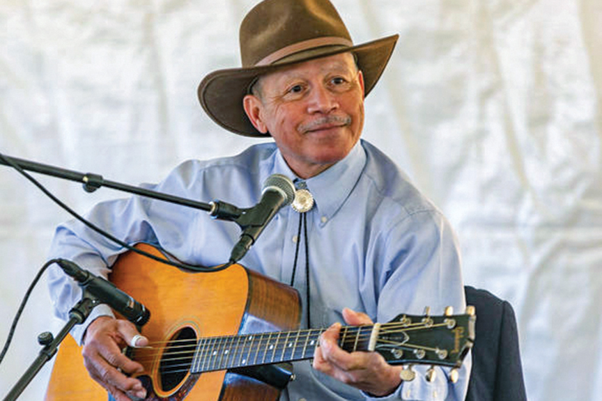 A Piedmont bluesman wearing a cowboy hat and bolo tie smiles as he plays an acoustic guitar into a microphone.