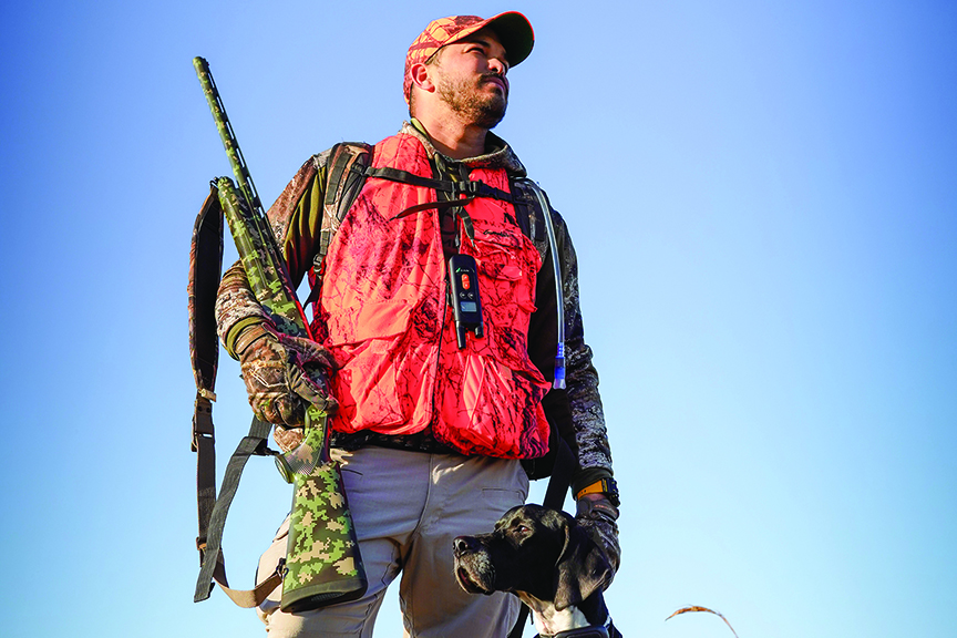 Hunter in orange vest and cap stands with rifle as his black dog sits at his feet