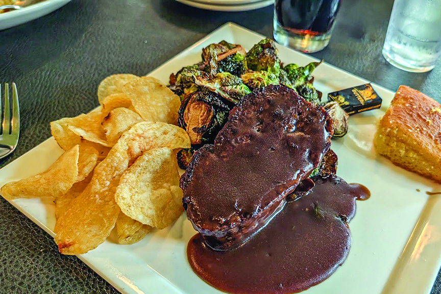 A square white plate contains meatloaf covered in gravy, kettle chips, and Brussels sprouts