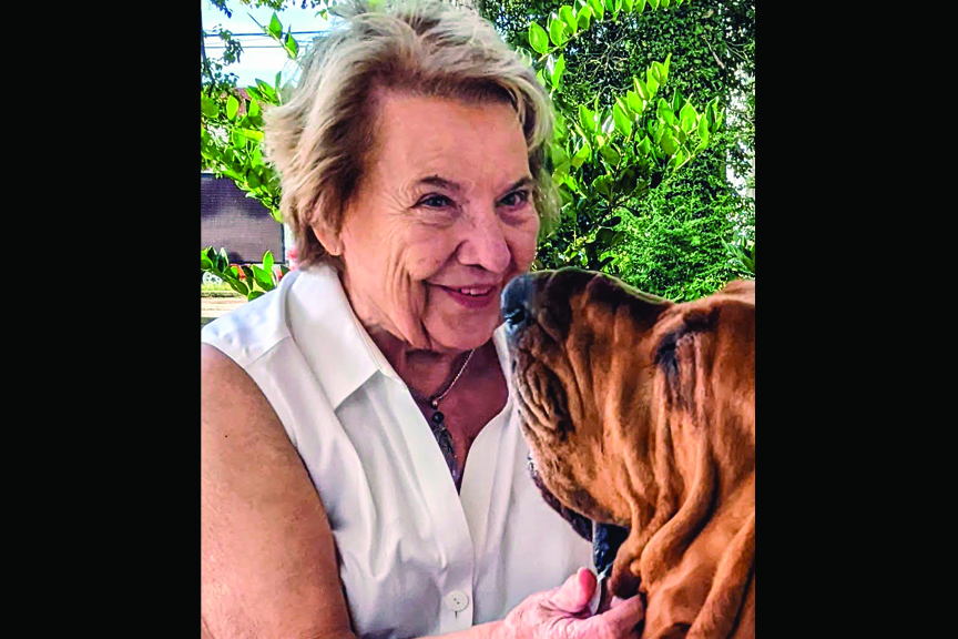 Polly, a smiling woman with short blonde hair, gently holding a reddish-brown hound dog looking closely at her face.