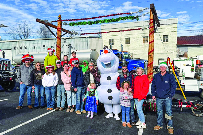 Large group of festive people, including one person in an Olaf costume, pose for a photo in front of floats during a holiday parade.